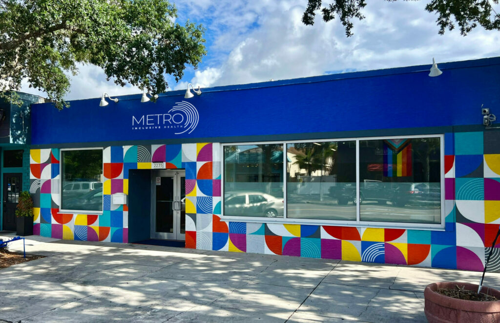 Front entrance of Metro Inclusive Health on Central Ave. in St. Pete, Florida. A colorful mural exterior with METRO branded colors including blue, red, teal, orange and purple. Above are blue skies and oak trees on both sides.